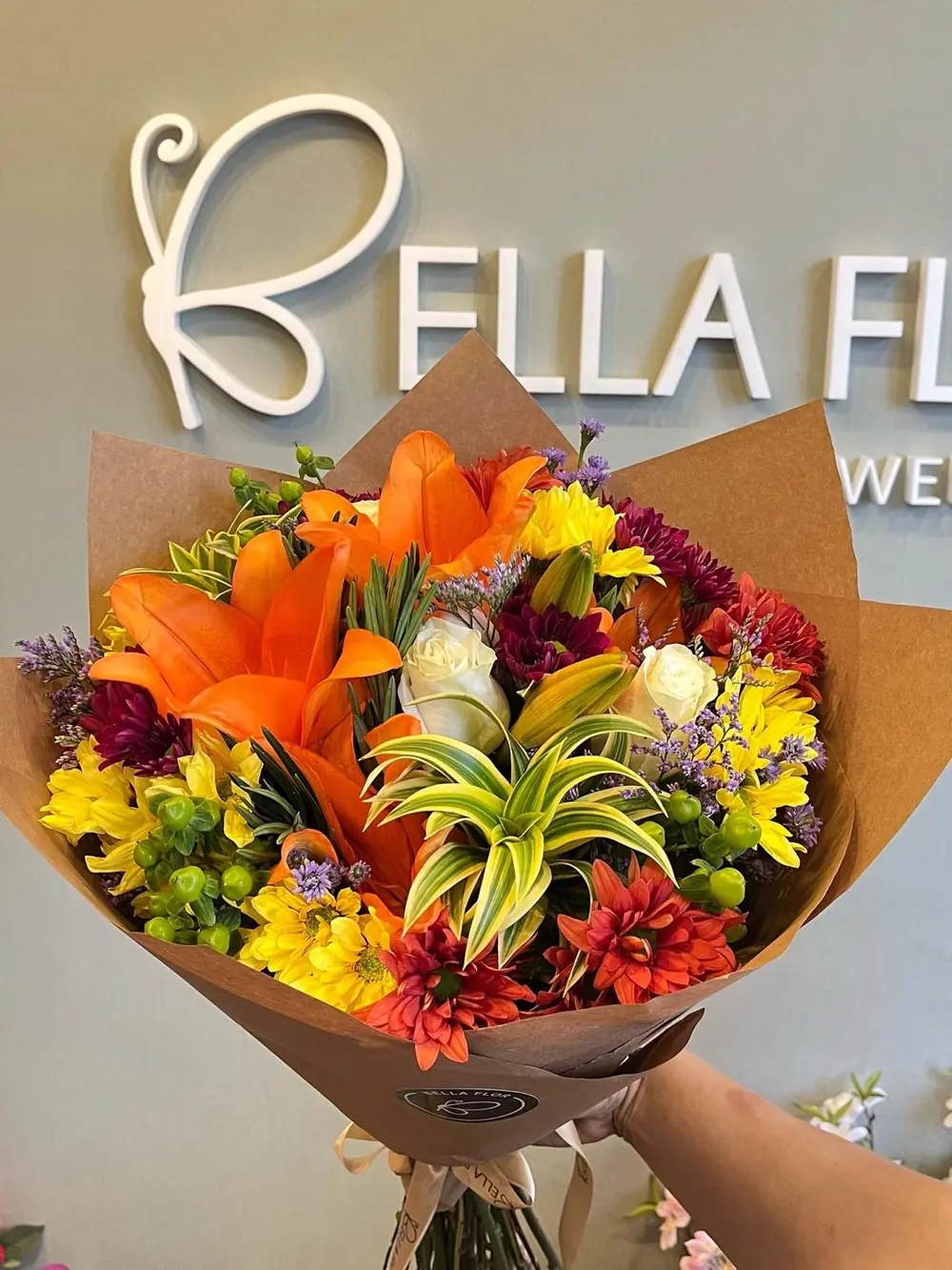 Ramo de lirios naranjas y flores de colores sostenido frente al cartel BELLA FLOR de la florería
