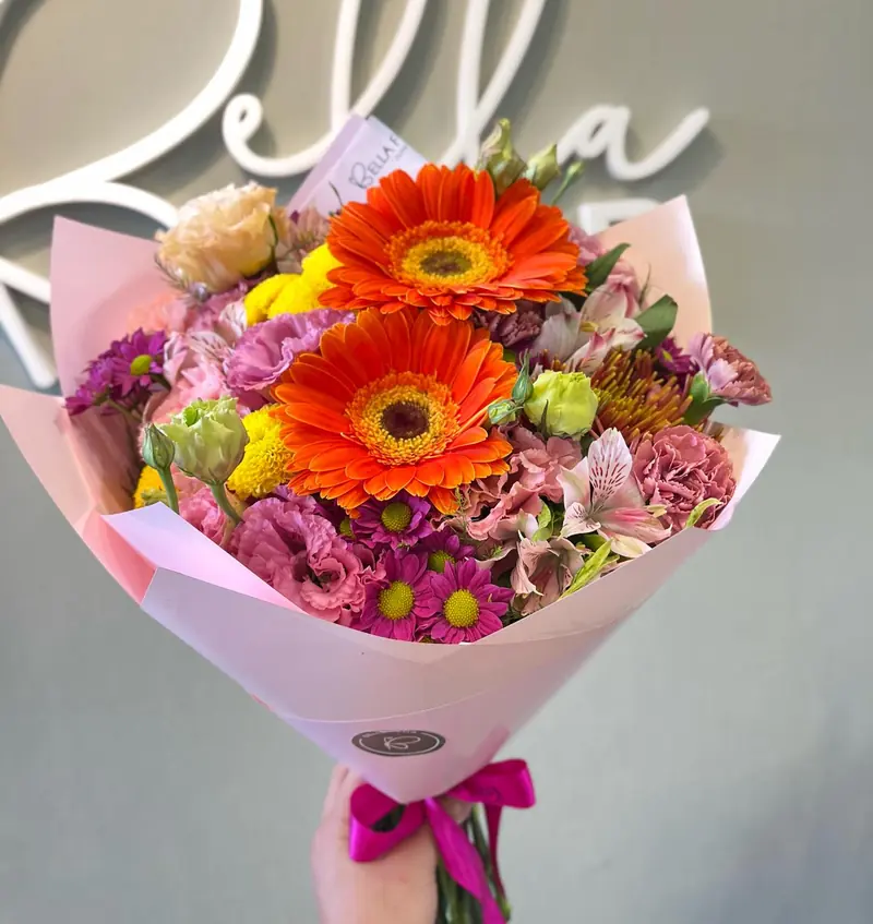 Ramo multicolor con gerberas naranjas en papel rosa, con el cartel Bella de fondo
